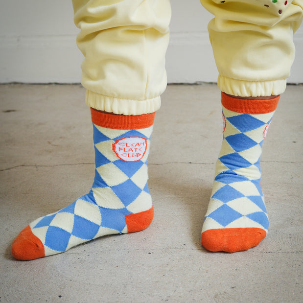 Kids Brrr Los Angeles Clean Plate Club socks featuring a yellow and blue diamond knit pattern with orange accents on the ankle, heel, and toe, and Clean Plate Club logos on both ankles. Styled with yellow Brrr sweatpants, part of the Brrr kidswear collection inspired by playful food-themed designs.