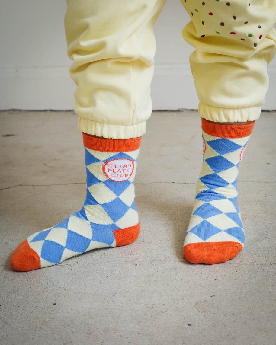 Kids Clean Plate Club socks with yellow and blue diamond knit pattern, orange accents on ankle, heel, and toe, featuring Clean Plate Club logos on both ankles, styled with Brrr yellow sweatpants