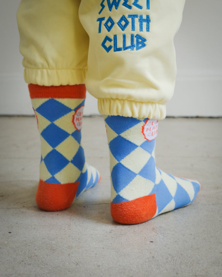 Back view of kids socks in yellow and blue diamond knit design with orange ankle, heel, and toe details, Clean Plate Club logos on both ankles, paired with Brrr yellow sweatpants