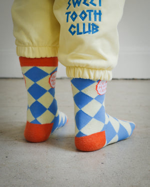 Back view of kids socks in yellow and blue diamond knit design with orange ankle, heel, and toe details, Clean Plate Club logos on both ankles, paired with Brrr yellow sweatpants