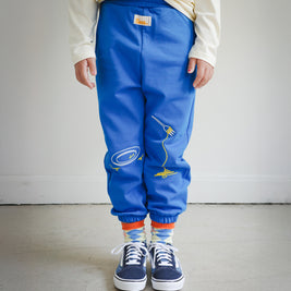 Kids blue sweatpants featuring Brrr main label below the front waistband, chain-stitched embroidery of a clean plate motif on the left front leg, and spaghetti noodles draped over a fork embroidered on the right front leg. Styled with sneakers for a playful look.