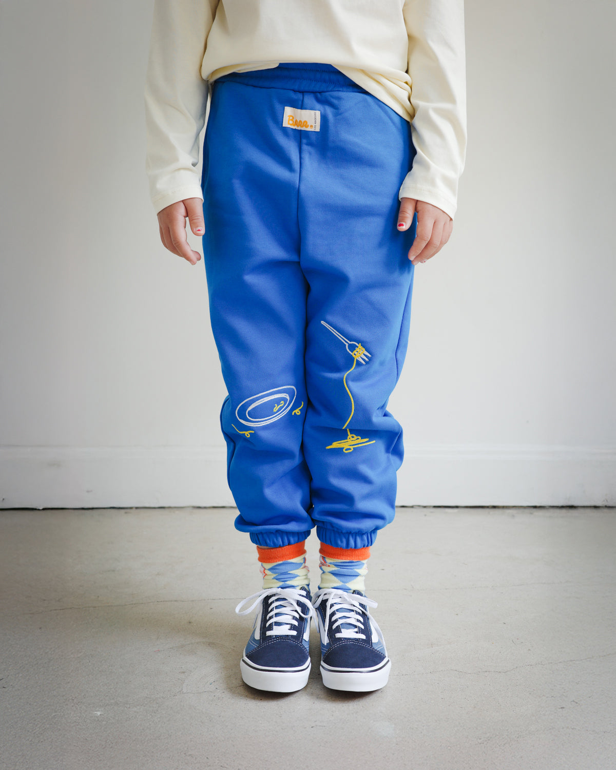 Kids blue sweatpants featuring Brrr main label below the front waistband, chain-stitched embroidery of a clean plate motif on the left front leg, and spaghetti noodles draped over a fork embroidered on the right front leg. Styled with sneakers for a playful look.