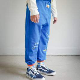 Side view of kids blue sweatpants with food-themed chain embroidery, showing clean plate design on the left leg and spaghetti fork motif on the right leg.