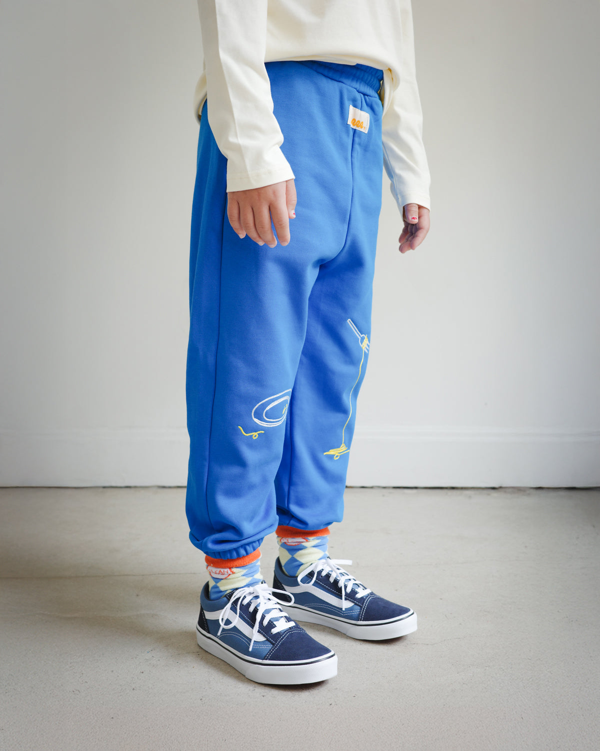 Side view of kids blue sweatpants with food-themed chain embroidery, showing clean plate design on the left leg and spaghetti fork motif on the right leg.