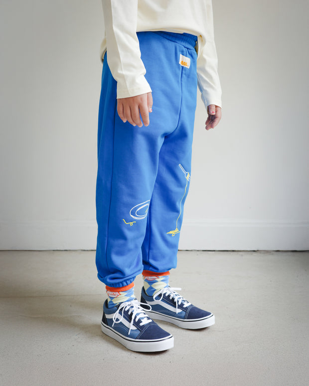 Side view of kids blue sweatpants with food-themed chain embroidery, showing clean plate design on the left leg and spaghetti fork motif on the right leg.