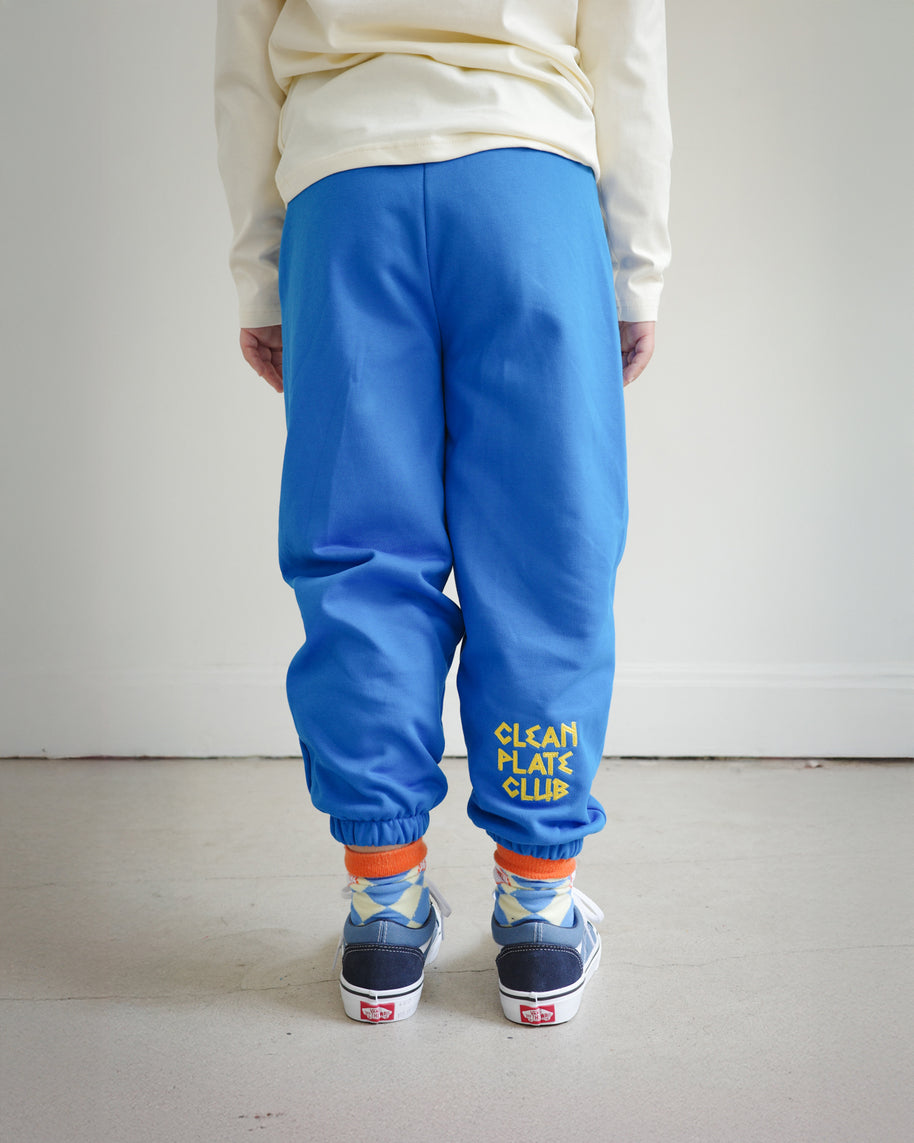 Back view of kids blue sweatpants with “Clean Plate Club” chain embroidery on the right back leg, elastic cuffs at the ankle.