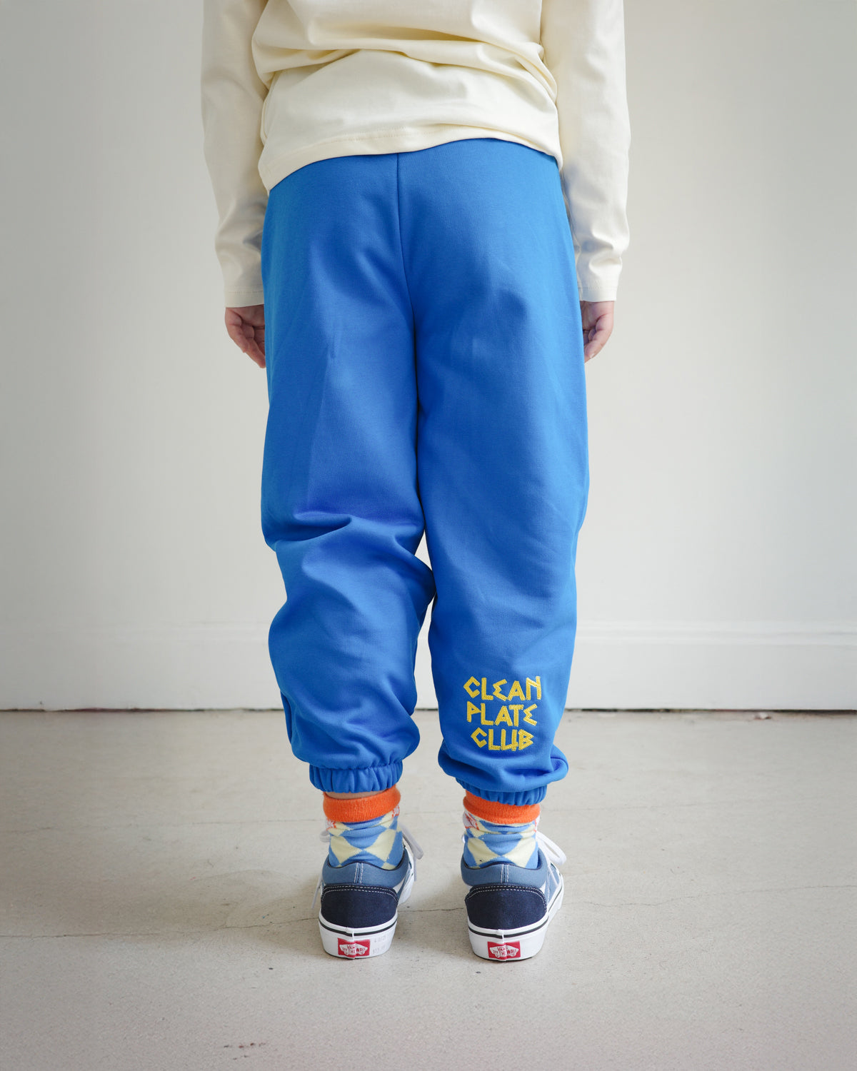 Back view of kids blue sweatpants with “Clean Plate Club” chain embroidery on the right back leg, elastic cuffs at the ankle.