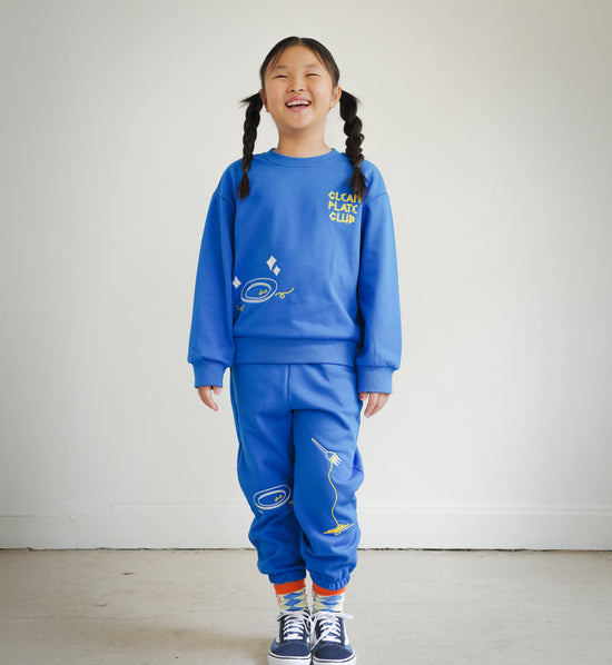Full body view of child wearing Brrr Los Angeles Clean Plate Club kids sweatshirt and matching sweatpants in bright blue, featuring playful chain-stitched embroidery of plate and spaghetti motifs. Part of the Brrr kidswear collection, designed for comfort, color, and fun food-inspired style.