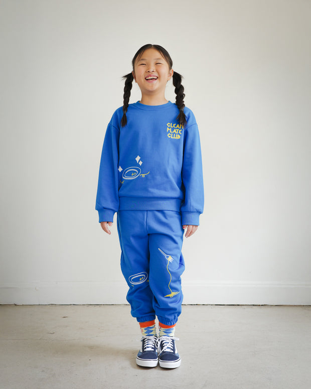 Full body view of child wearing Brrr Los Angeles Clean Plate Club kids sweatshirt and matching sweatpants in bright blue, featuring playful chain-stitched embroidery of plate and spaghetti motifs. Part of the Brrr kidswear collection, designed for comfort, color, and fun food-inspired style.