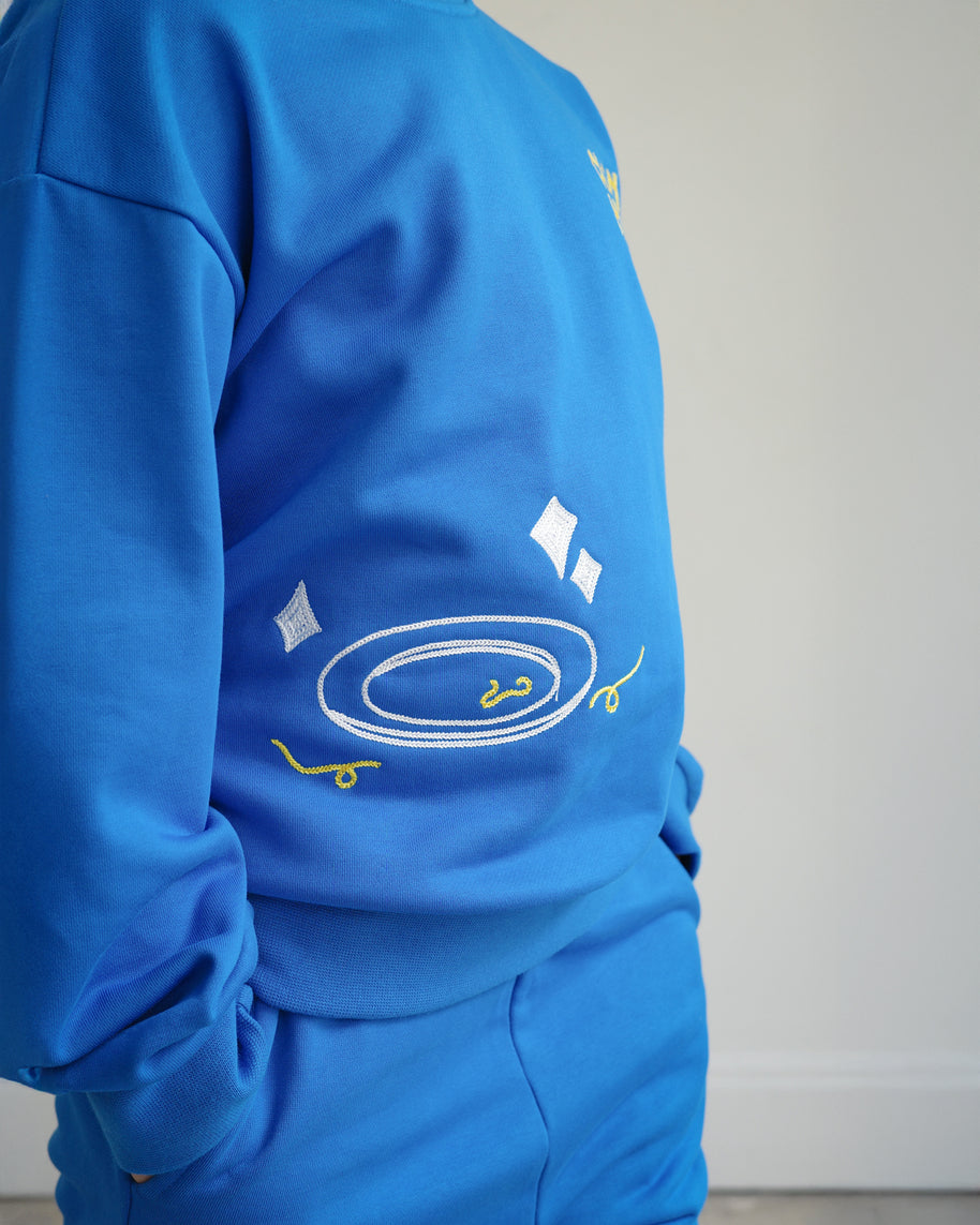 Close-up of Brrr kids blue sweatshirt from the Clean Plate Club collection, featuring chain-stitched embroidery of a clean white plate with playful yellow spaghetti details on the front. Made with a soft cotton-polyester blend, designed for comfort and durability in children’s everyday wear.
