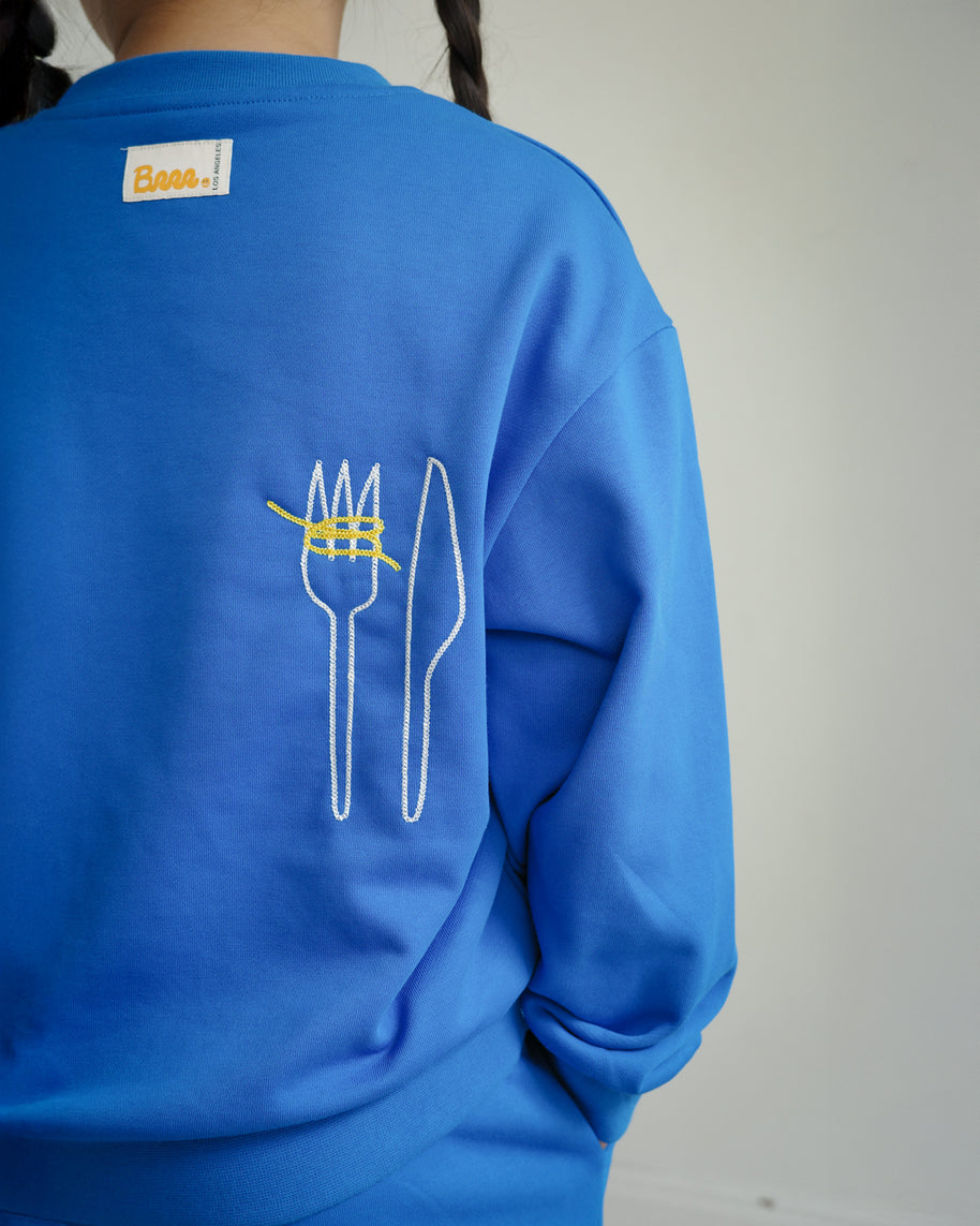 Back view of Brrr kids blue sweatshirt from the Clean Plate Club collection, featuring chain-stitched embroidery of a fork and knife with playful yellow spaghetti detail. The sweatshirt also includes the signature Brrr main label stitched at the back neckline, designed in a cotton-polyester blend for comfort and durability in children’s fashion.