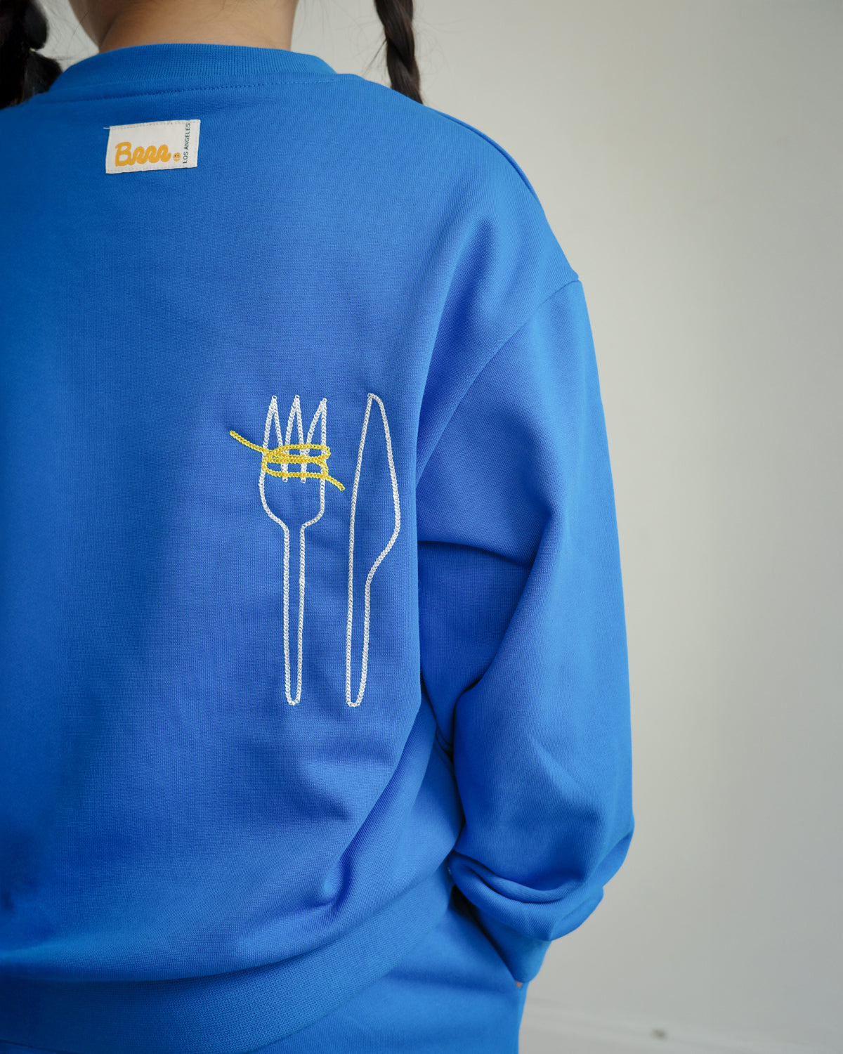 Back view of Brrr kids blue sweatshirt from the Clean Plate Club collection, featuring chain-stitched embroidery of a fork and knife with playful yellow spaghetti detail. The sweatshirt also includes the signature Brrr main label stitched at the back neckline, designed in a cotton-polyester blend for comfort and durability in children’s fashion.
