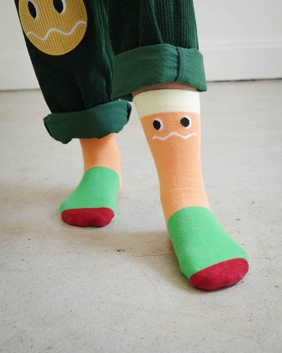 Kids Brrr Face color block socks in yellow, orange, green, and burgundy knit sections, featuring the Brrr face logo on the orange ankle, styled with green corduroy overalls