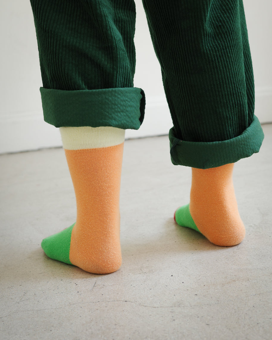 Back view of kids color block socks with four-tone knit design in yellow, orange, green, and burgundy, with Brrr face logo detail on the ankle, paired with rolled-up green overalls