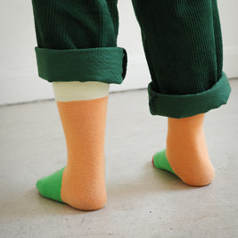 Back view of kids color block socks with four-tone knit design in yellow, orange, green, and burgundy, with Brrr face logo detail on the ankle, paired with rolled-up green overalls