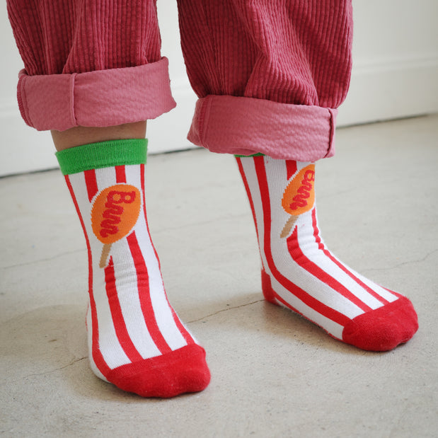 Kids Brrr Los Angeles corn dog striped socks featuring vertical red and white knit stripes, a green ankle cuff, and a corn dog graphic with a Brrr logo–shaped ketchup detail. Styled with rose corduroy pants, part of the Brrr kidswear collection inspired by playful food-themed design.