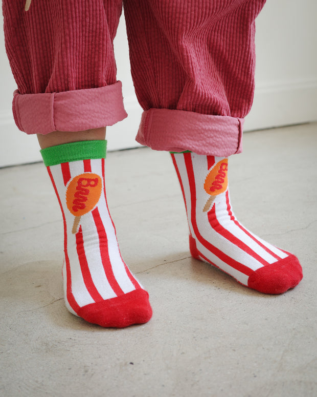 Kids corn dog striped socks with vertical red and white knit stripes, green ankle cuff, and corn dog graphic with Brrr logo-shaped ketchup detail, styled with rose corduroy pants