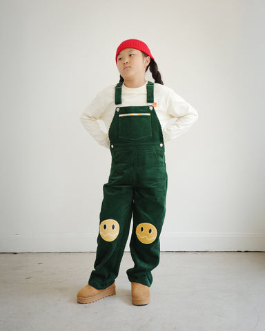 Full body view of child wearing Brrr kids green corduroy overalls with yellow smiley face knee patches, part of the Brrr Los Angeles kids clothing collection.