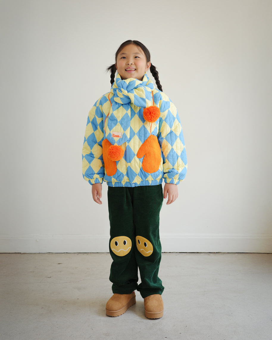 Full body view of child wearing matching blue and yellow diamond pattern quilted puff scarf with orange pompoms, paired with Brrr quilted jacket and knee patch green corduroy overalls for a coordinated look