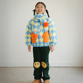 Full body view of child wearing matching blue and yellow diamond pattern quilted puff scarf with orange pompoms, paired with Brrr quilted jacket and knee patch green corduroy overalls for a coordinated look