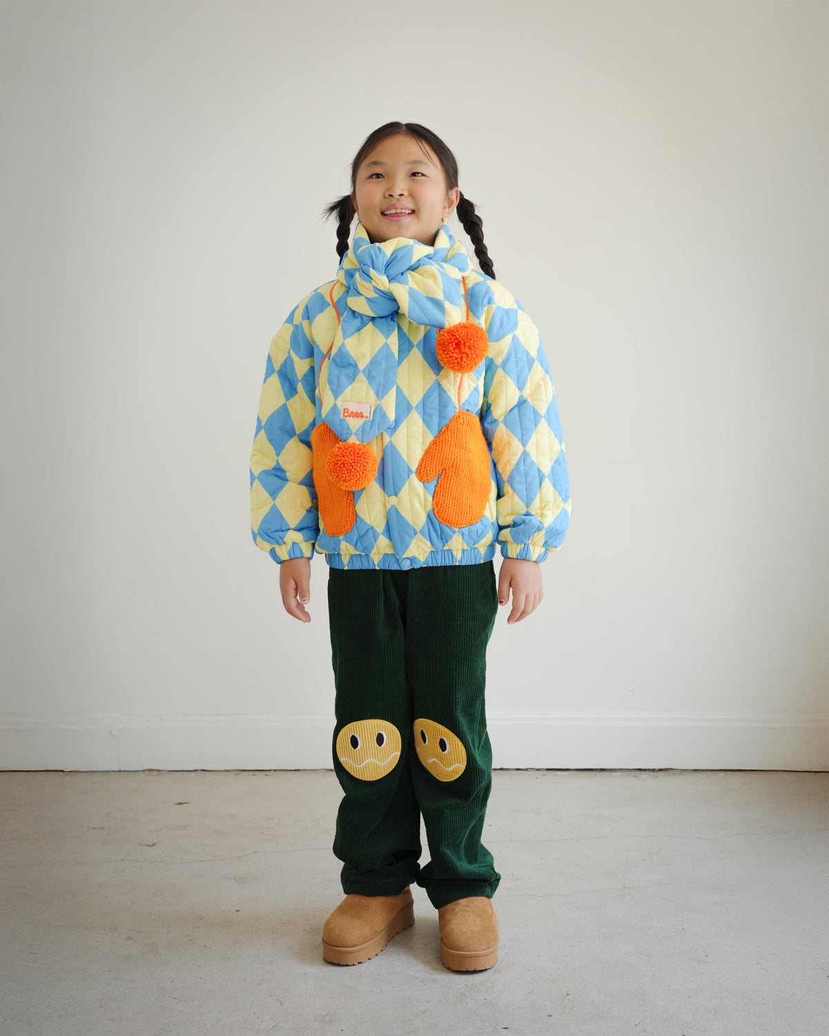 Full body view of child wearing matching blue and yellow diamond pattern quilted puff scarf with orange pompoms, paired with Brrr quilted jacket and knee patch green corduroy overalls for a coordinated look