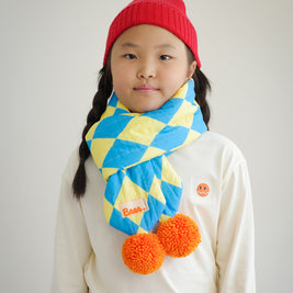 Front view of kids quilted puff scarf with diamond pattern fabric and playful orange pompoms, styled with ivory long sleeve shirt