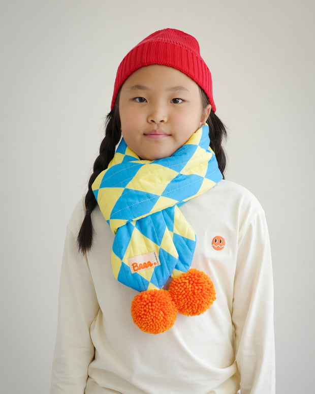 Front view of kids quilted puff scarf with diamond pattern fabric and playful orange pompoms, styled with ivory long sleeve shirt