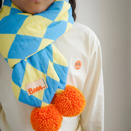 Close-up of children’s quilted puff scarf featuring blue and yellow diamond pattern fabric and two large orange pompoms
