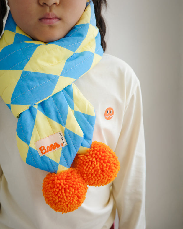 Close-up of children’s quilted puff scarf featuring blue and yellow diamond pattern fabric and two large orange pompoms