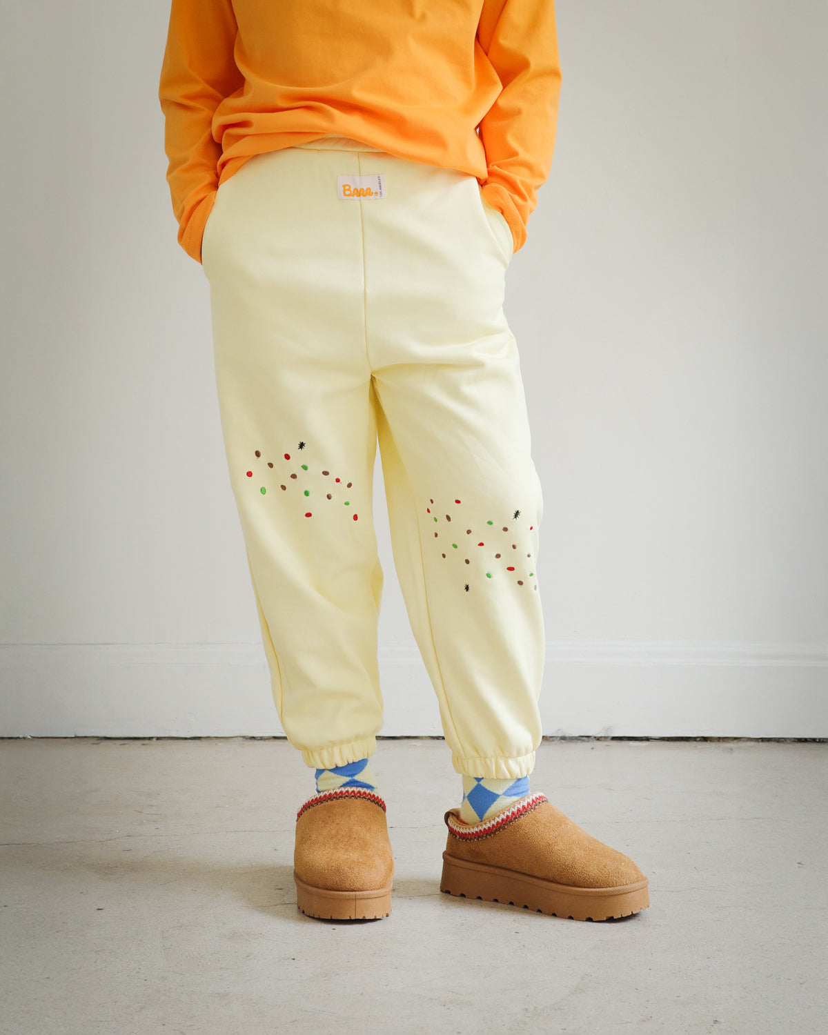 Kids yellow sweatpants from the Brrr Sweet Tooth Club collection, featuring playful food-themed chain embroidery. The front thigh is decorated with cookie crumb embroidery and an ant motif for a whimsical detail. Made with a soft cotton blend and elastic waistband for comfort in children’s everyday wear.