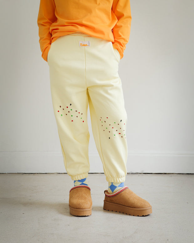 Kids yellow sweatpants from the Brrr Sweet Tooth Club collection, featuring playful food-themed chain embroidery. The front thigh is decorated with cookie crumb embroidery and an ant motif for a whimsical detail. Made with a soft cotton blend and elastic waistband for comfort in children’s everyday wear.