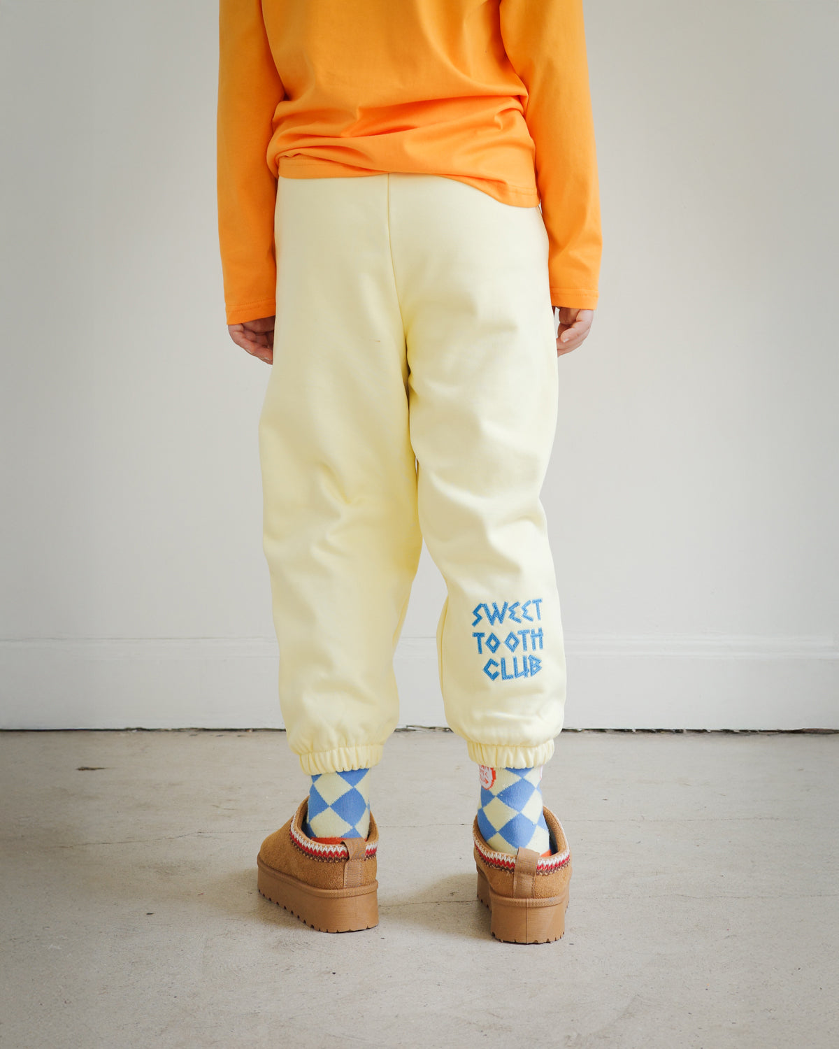 Back view of kids yellow sweatpants from Brrr, showcasing bold chain-stitched “Sweet Tooth Club” text embroidery along the back leg. Made in a medium-weight cotton blend, perfect for cooler weather children’s fashion.