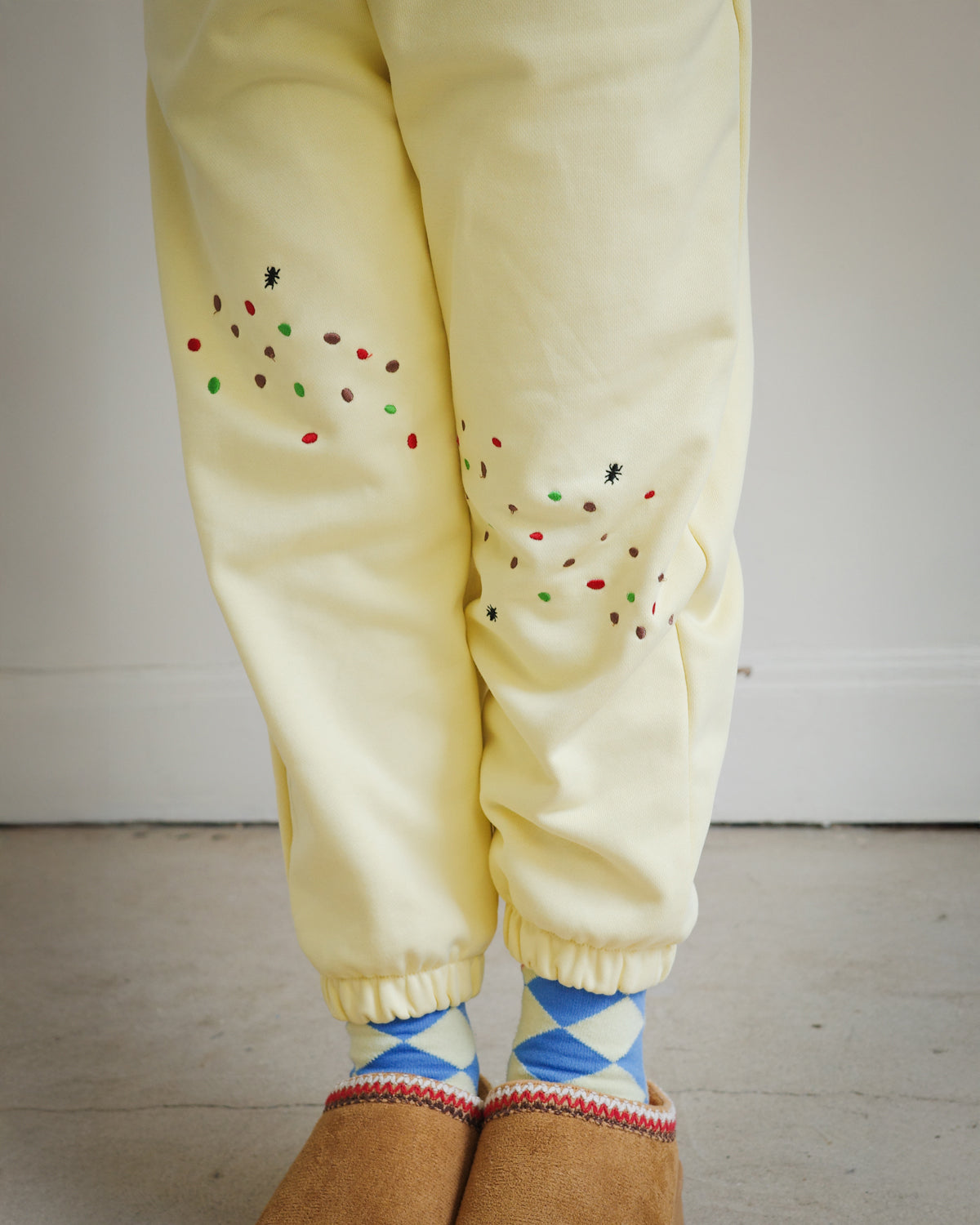 Close-up of yellow kids sweatpants with food-themed embroidery, highlighting cookie crumb and ant motifs at the front thigh and Sweet Tooth Club lettering at the back. Part of Brrr’s playful kidswear collection designed with comfort, stretch, and whimsical embroidered details.