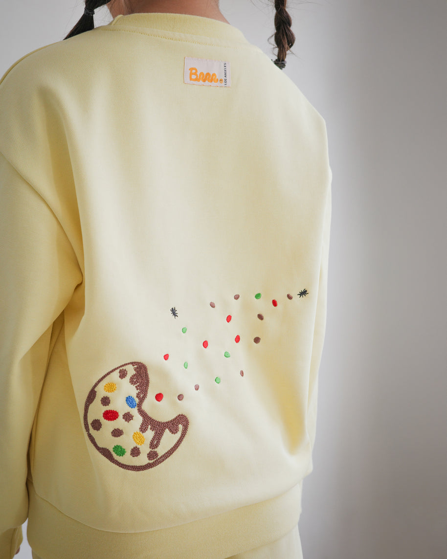 Back view of Brrr kids yellow sweatshirt from the Sweet Tooth Club collection, featuring chain-stitched embroidery of a bitten cookie with colorful crumbs scattered across the fabric, accented by tiny ant motifs. The sweatshirt also includes the signature Brrr main label at the back neckline, combining playful food-inspired details with cozy kidswear design.