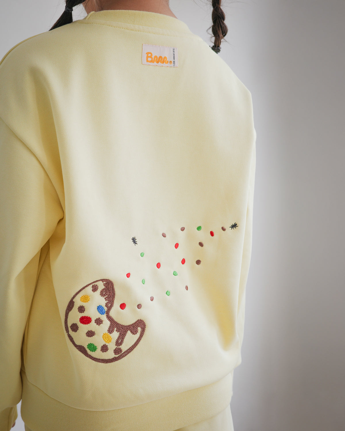 Back view of Brrr kids yellow sweatshirt from the Sweet Tooth Club collection, featuring chain-stitched embroidery of a bitten cookie with colorful crumbs scattered across the fabric, accented by tiny ant motifs. The sweatshirt also includes the signature Brrr main label at the back neckline, combining playful food-inspired details with cozy kidswear design.