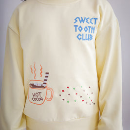 Close-up of yellow kids sweatshirt highlighting chain-stitched embroidery details: “Sweet Tooth Club” text, hot cocoa cup, cookie crumbs and ant motifs on the front