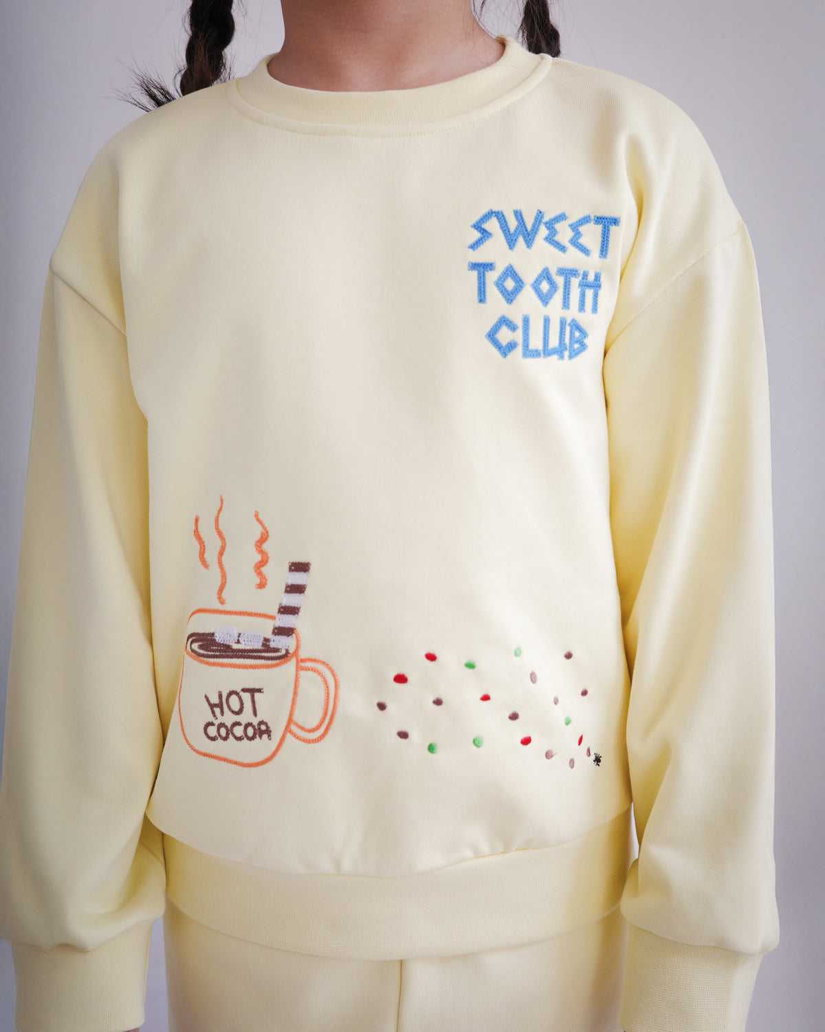 Close-up of yellow kids sweatshirt highlighting chain-stitched embroidery details: “Sweet Tooth Club” text, hot cocoa cup, cookie crumbs and ant motifs on the front