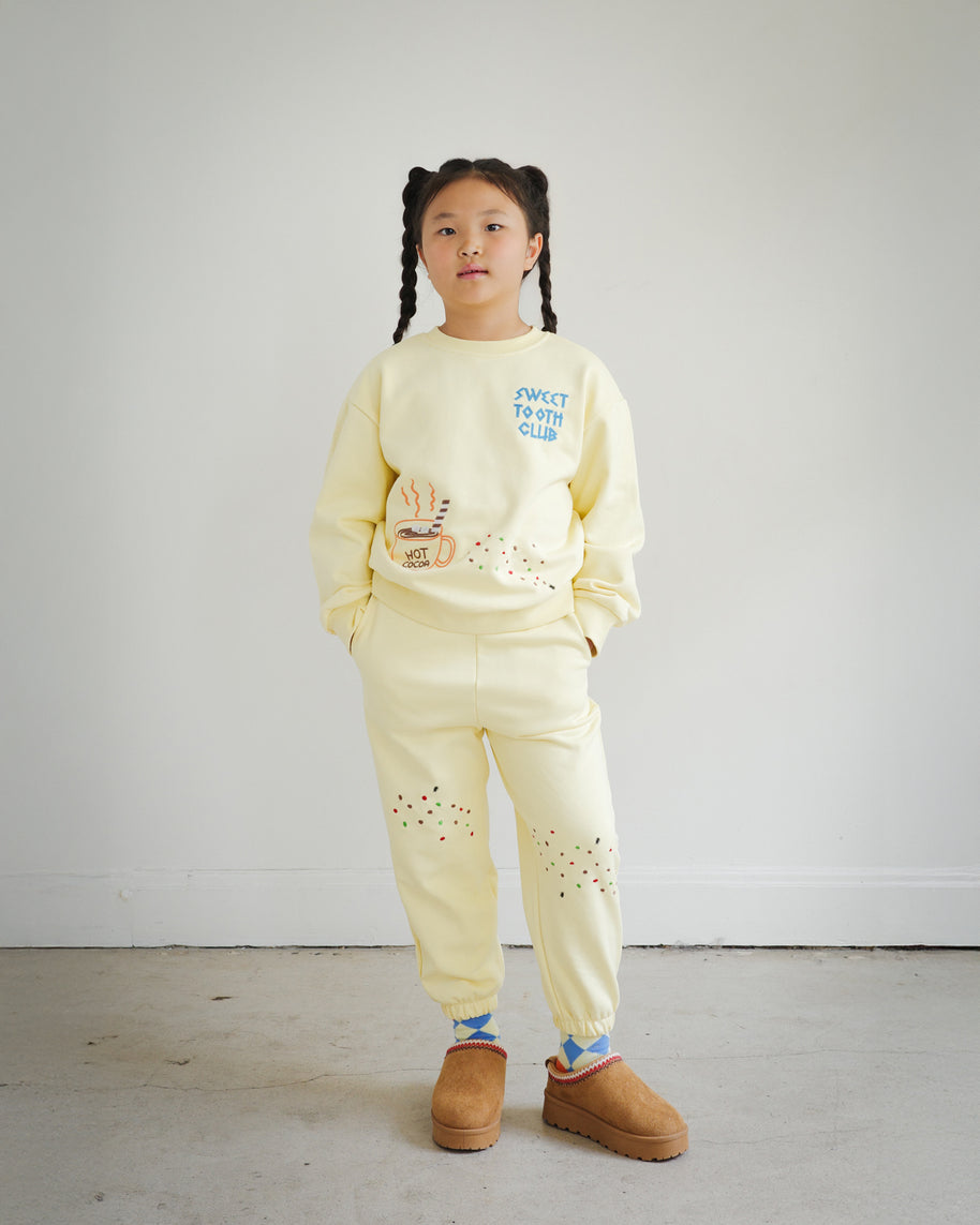 Full outfit view of Brrr kids yellow sweatshirt and sweatpants set from the Sweet Tooth Club collection. The sweatshirt features chain-stitched embroidery of “Sweet Tooth Club” text, a hot cocoa cup, cookie crumbs and ant motifs on the front, while the matching sweatpants are decorated with playful cookie crumb and ant embroidery on the thighs. Made with a cotton-polyester blend, this coordinated kidswear set combines comfort, durability, and whimsical food-themed design.