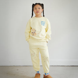 Full outfit view of Brrr kids yellow sweatshirt and sweatpants set from the Sweet Tooth Club collection. The sweatshirt features chain-stitched embroidery of “Sweet Tooth Club” text, a hot cocoa cup, cookie crumbs and ant motifs on the front, while the matching sweatpants are decorated with playful cookie crumb and ant embroidery on the thighs. Made with a cotton-polyester blend, this coordinated kidswear set combines comfort, durability, and whimsical food-themed design.