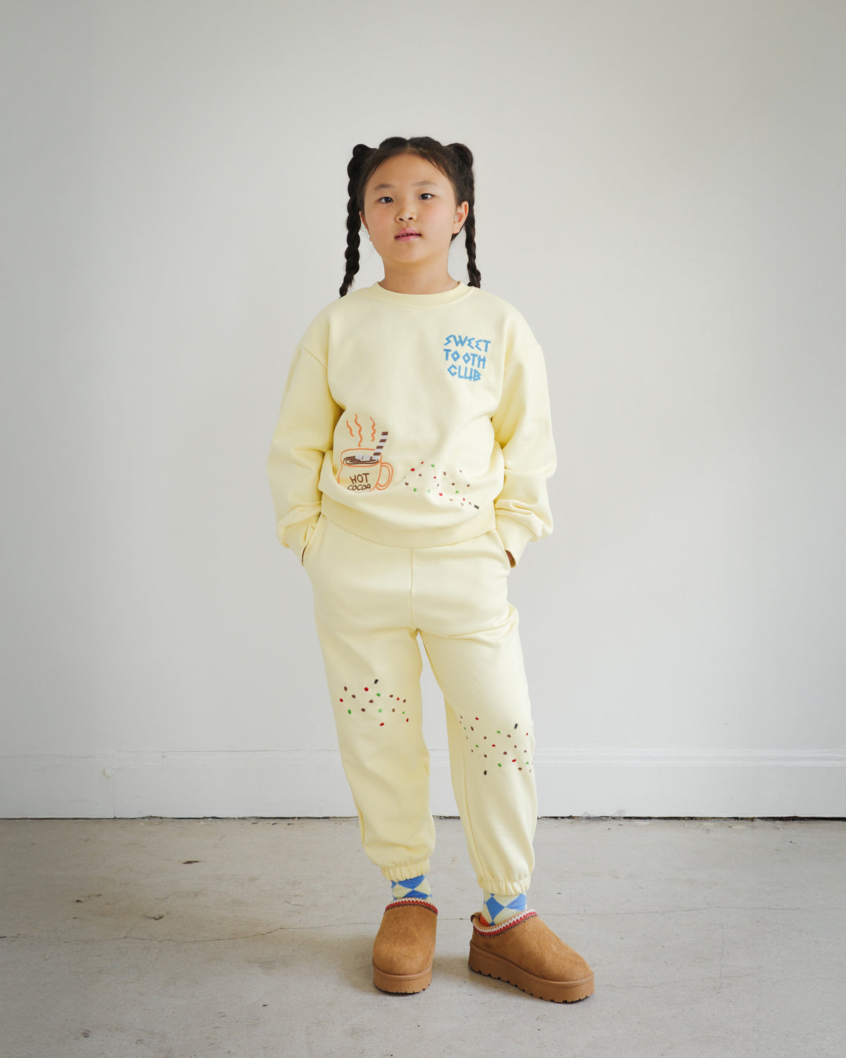 Full outfit view of Brrr kids yellow sweatshirt and sweatpants set from the Sweet Tooth Club collection. The sweatshirt features chain-stitched embroidery of “Sweet Tooth Club” text, a hot cocoa cup, cookie crumbs and ant motifs on the front, while the matching sweatpants are decorated with playful cookie crumb and ant embroidery on the thighs. Made with a cotton-polyester blend, this coordinated kidswear set combines comfort, durability, and whimsical food-themed design.