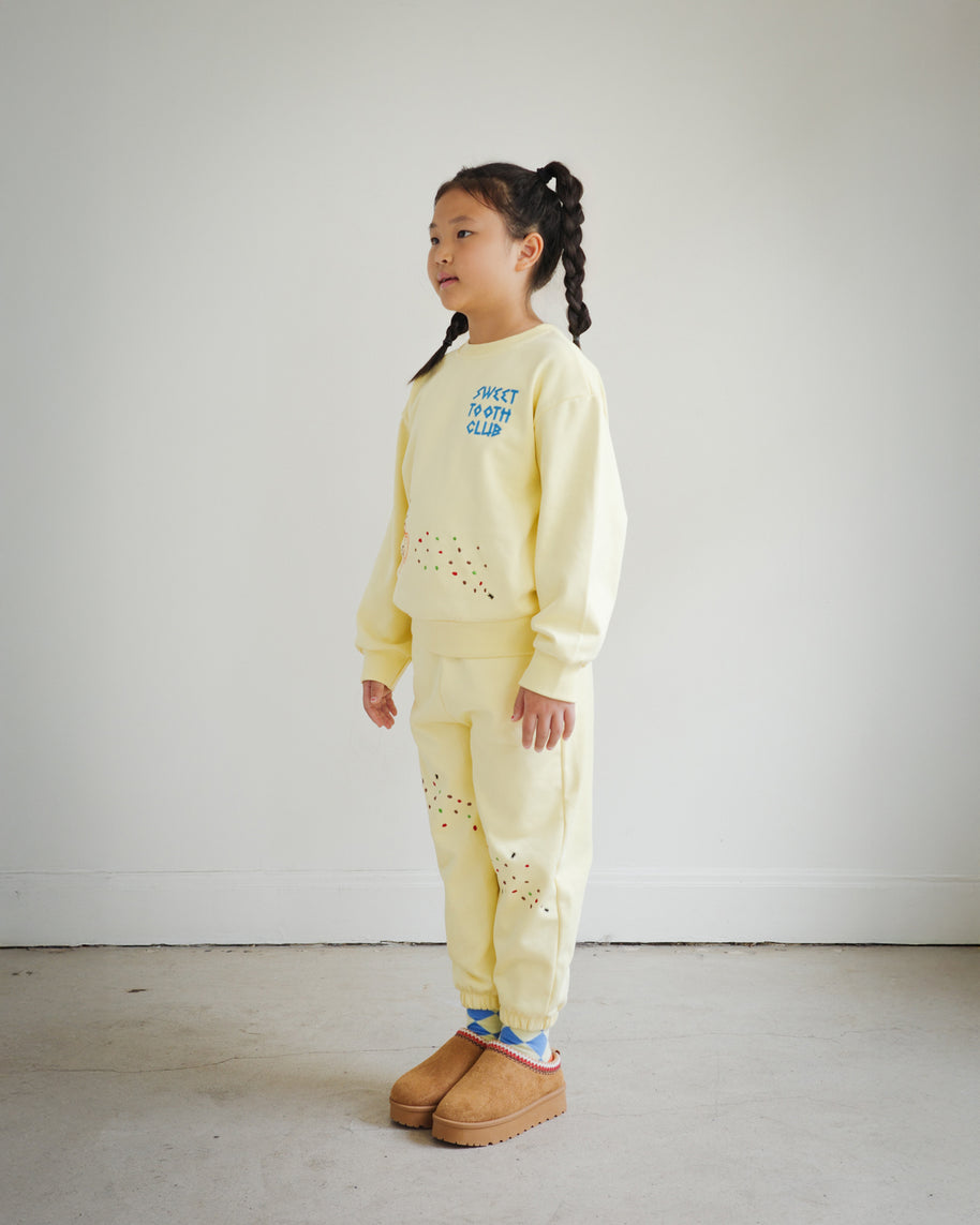 Side view of a child wearing the Brrr kids yellow Sweet Tooth Club sweatshirt and sweatpants set. The sweatshirt features blue “Sweet Tooth Club” text embroidery and playful cookie crumb and ant motifs on the front, while the matching sweatpants have food-themed embroidery details on both legs. Cozy cotton-polyester blend kidswear designed for comfort and style.