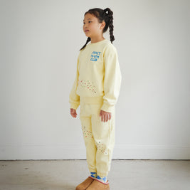 Side view of a child wearing the Brrr kids yellow Sweet Tooth Club sweatshirt and sweatpants set. The sweatshirt features blue “Sweet Tooth Club” text embroidery and playful cookie crumb and ant motifs on the front, while the matching sweatpants have food-themed embroidery details on both legs. Cozy cotton-polyester blend kidswear designed for comfort and style.