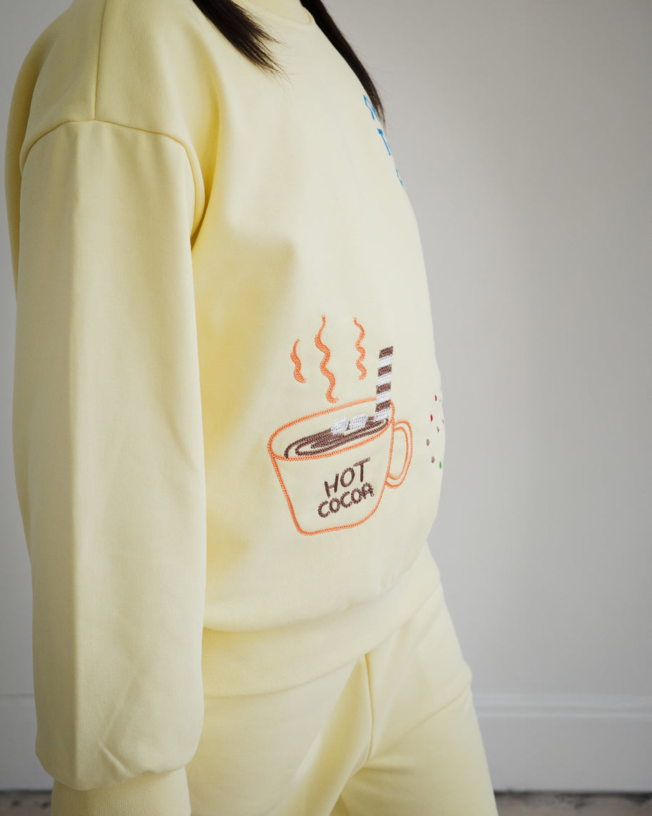 Close-up of Brrr kids yellow sweatshirt from the Sweet Tooth Club collection, featuring chain-stitched embroidery of a hot cocoa cup with steam and marshmallows. Designed in a soft cotton-polyester blend, this playful food-themed detail adds a cozy and whimsical touch to children’s fashion.