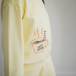 Close-up of Brrr kids yellow sweatshirt from the Sweet Tooth Club collection, featuring chain-stitched embroidery of a hot cocoa cup with steam and marshmallows. Designed in a soft cotton-polyester blend, this playful food-themed detail adds a cozy and whimsical touch to children’s fashion.