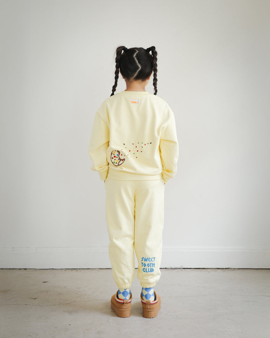 Back view of a child wearing the Brrr kids yellow Sweet Tooth Club sweatshirt and sweatpants set. The sweatshirt features playful cookie crumb embroidery on the back with colorful sprinkles and ants, while the pants showcase bold blue “Sweet Tooth Club” text embroidery on the back leg. The outfit highlights Brrr’s food-themed kidswear design with fun and cozy style.