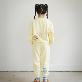Back view of a child wearing the Brrr kids yellow Sweet Tooth Club sweatshirt and sweatpants set. The sweatshirt features playful cookie crumb embroidery on the back with colorful sprinkles and ants, while the pants showcase bold blue “Sweet Tooth Club” text embroidery on the back leg. The outfit highlights Brrr’s food-themed kidswear design with fun and cozy style.