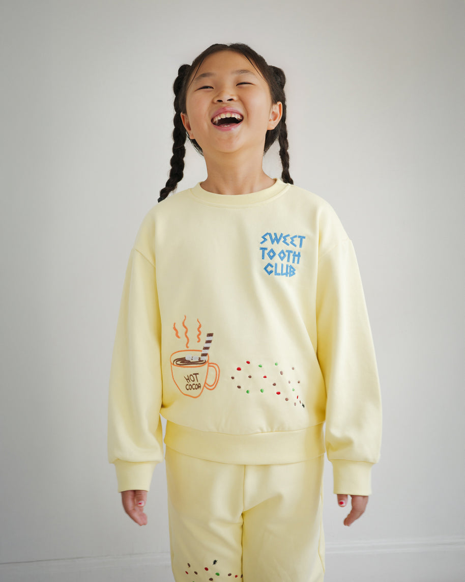 Kids yellow sweatshirt from the Brrr Sweet Tooth Club collection, featuring playful food-themed chain embroidery. The front showcases “Sweet Tooth Club” text embroidery with whimsical motifs of hot cocoa, cookie crumbs and ants, designed for a fun and cozy children’s fashion look.