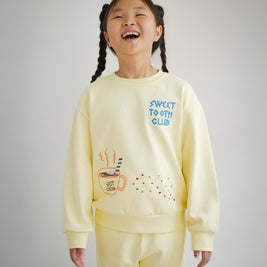 Kids yellow sweatshirt from the Brrr Sweet Tooth Club collection, featuring playful food-themed chain embroidery. The front showcases “Sweet Tooth Club” text embroidery with whimsical motifs of hot cocoa, cookie crumbs and ants, designed for a fun and cozy children’s fashion look.