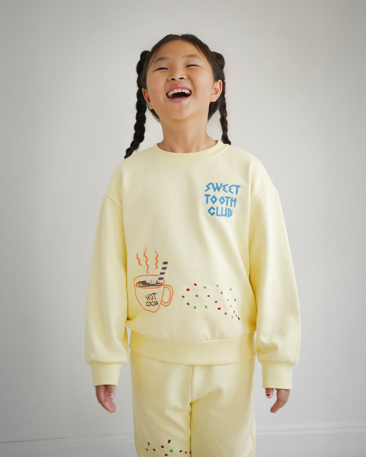 Kids yellow sweatshirt from the Brrr Sweet Tooth Club collection, featuring playful food-themed chain embroidery. The front showcases “Sweet Tooth Club” text embroidery with whimsical motifs of hot cocoa, cookie crumbs and ants, designed for a fun and cozy children’s fashion look.