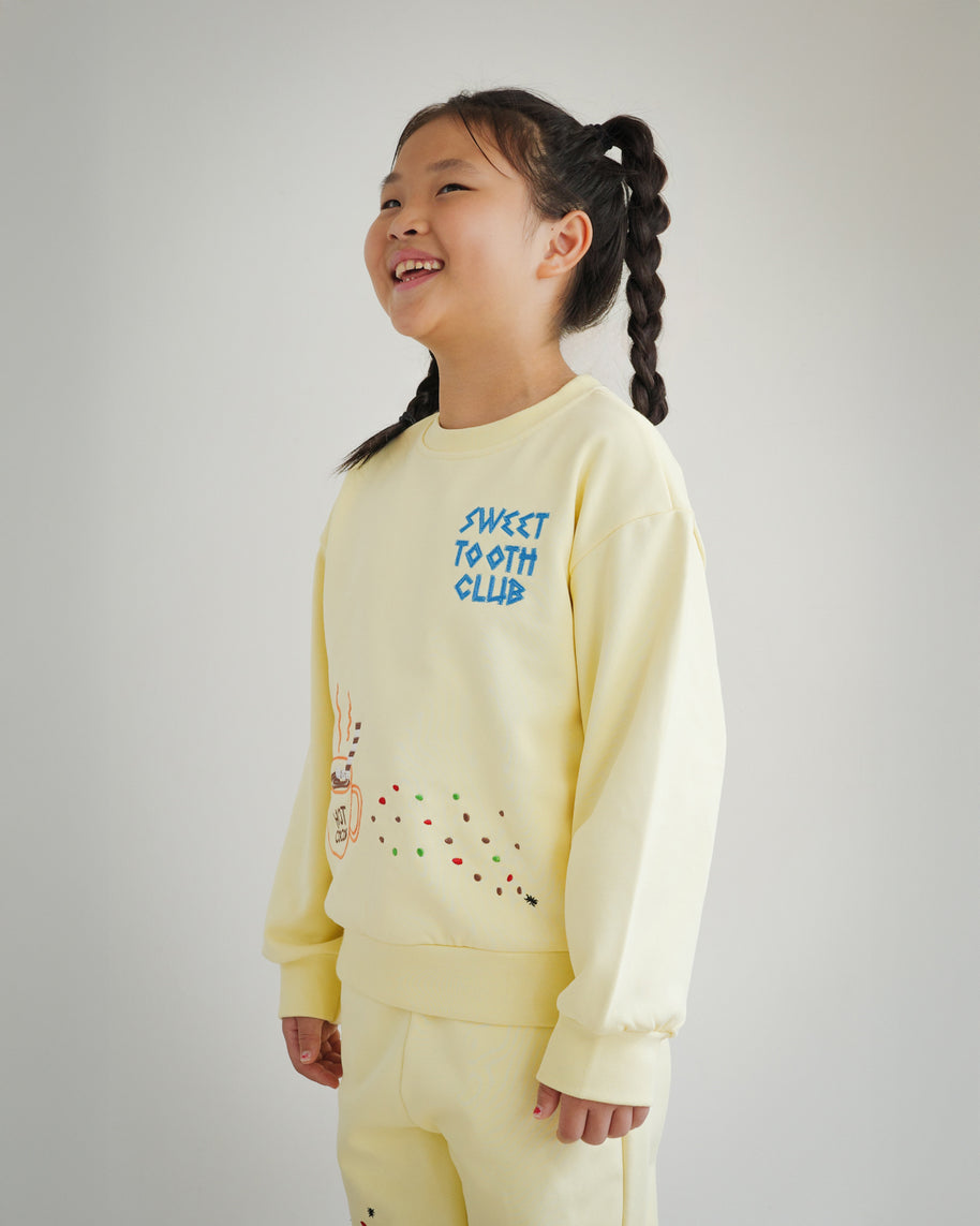 Side view of Brrr kids yellow sweatshirt with Sweet Tooth Club embroidery. Details include hot cocoa, cookie crumb and ant chain-stitched motifs on the front, made with a medium-weight cotton blend for comfort and durability in kidswear.